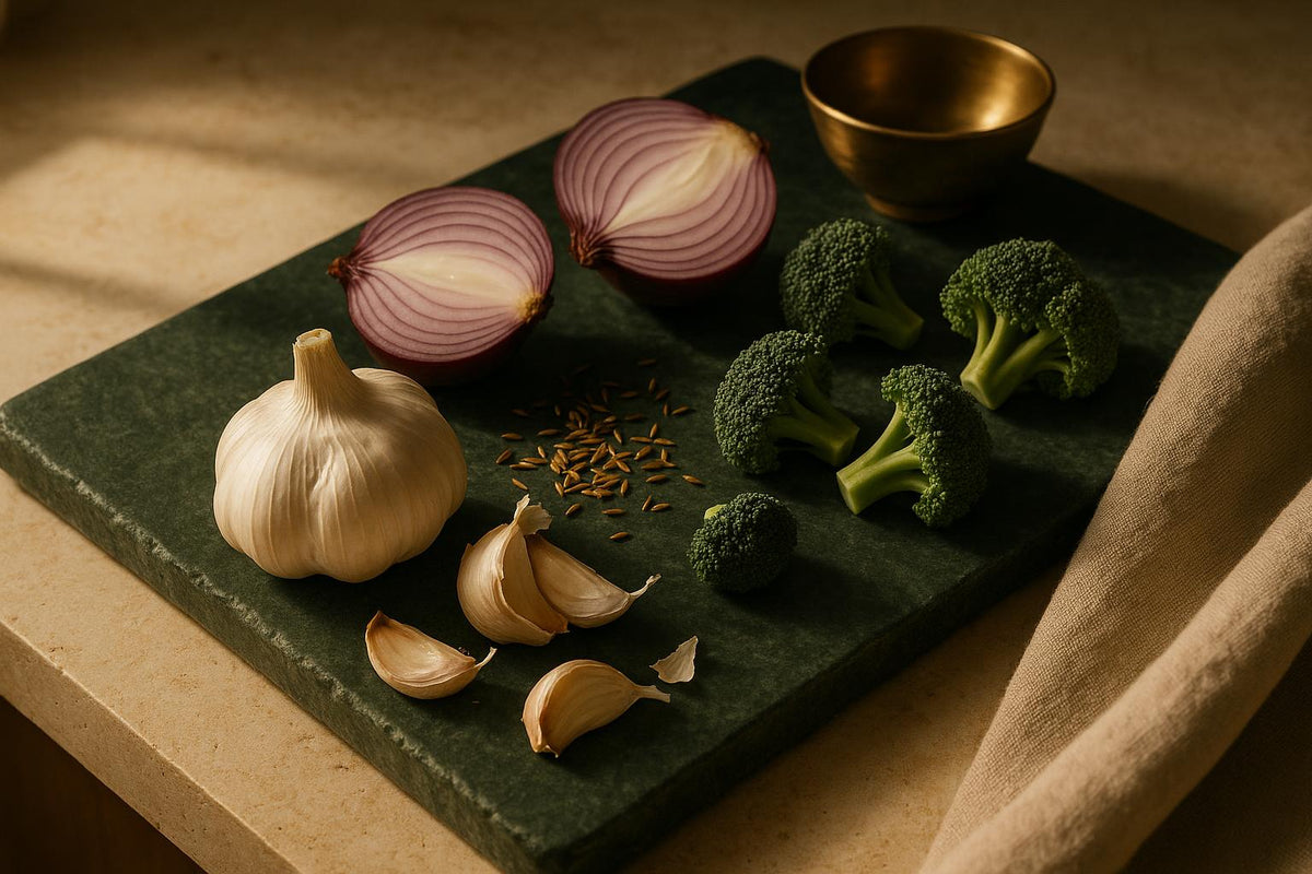 Whole garlic bulbs, halved red onions and broccoli florets arranged on a dark stone surface in warm side light